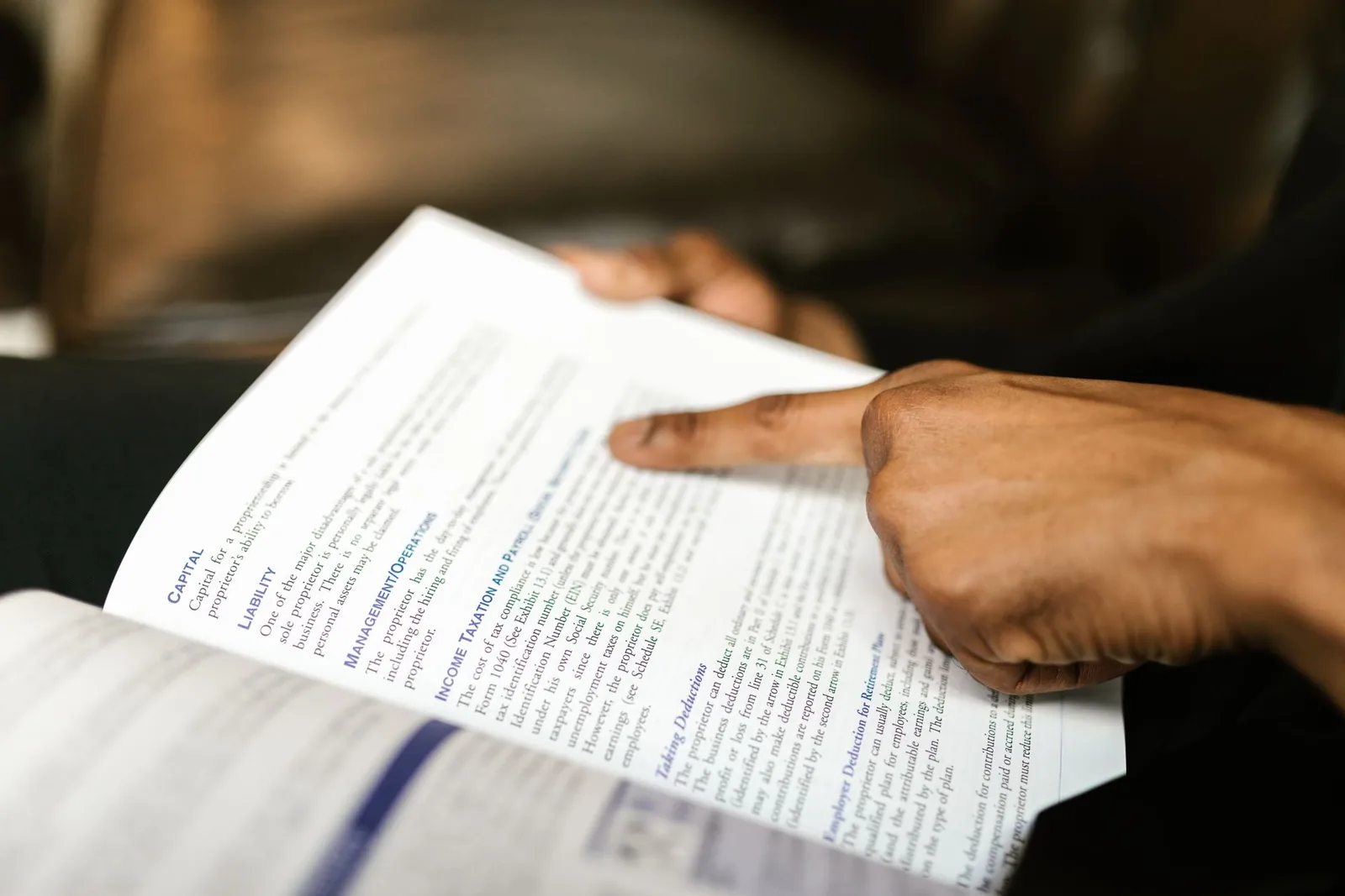 A finger pointing to a line of printed text in an open book, with the rest of the page out of focus