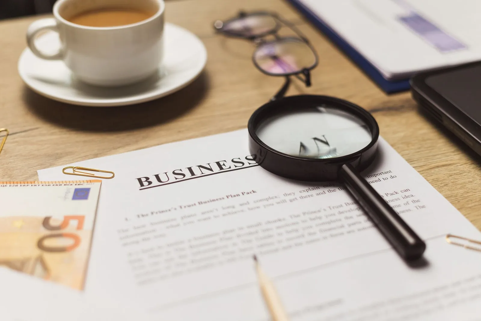 A magnifying glass resting on a printed business plan beside a cup of coffee, standing in for looking closely at what assistants say about a business.